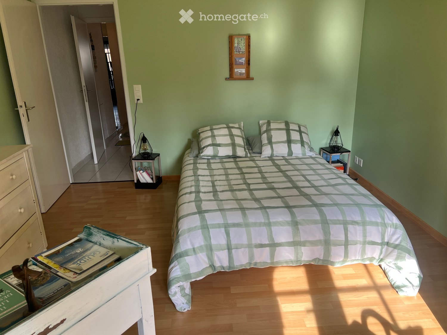 Bedroom. Cozy bedroom with a neatly made bed, nightstands, a small table with magazines, and a dresser. Light green walls and wooden flooring.