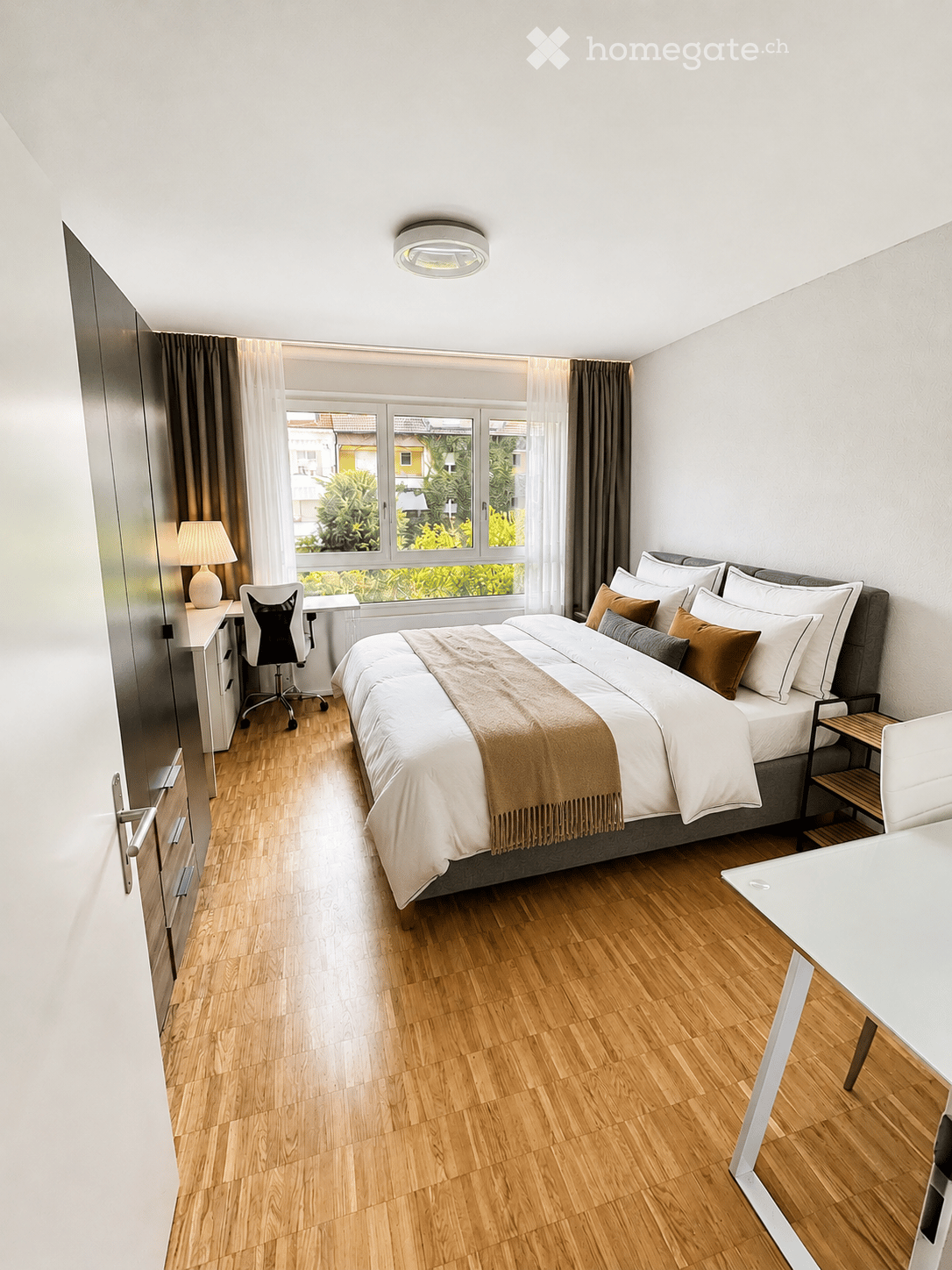 Modern bedroom with a large bed, wooden flooring, a desk with a chair, and a window with curtains.