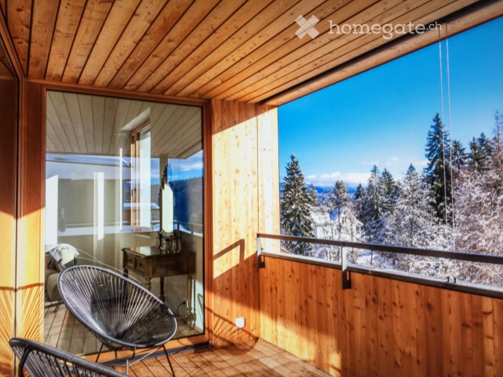 Wooden balcony with large windows offering a scenic mountain view, featuring a modern chair and a small table.