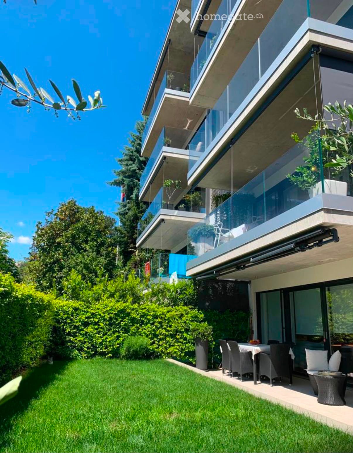Modern multi-story building with large glass balconies, surrounded by a well-maintained garden with green lawn and shrubs. Outdoor seating area includes a table, chairs, and a sofa.