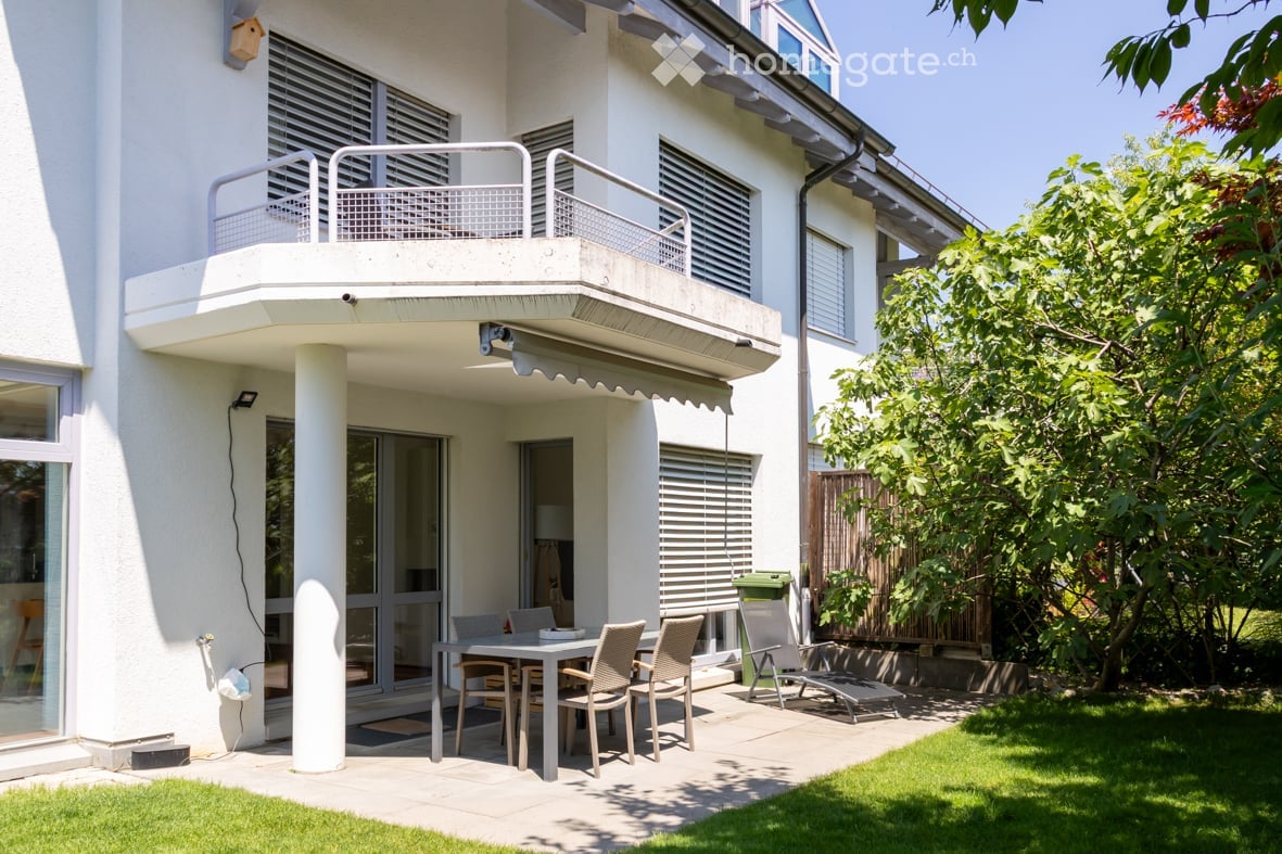 Balcony area with a table and chairs, surrounded by greenery and a modern building with large windows and shutters.