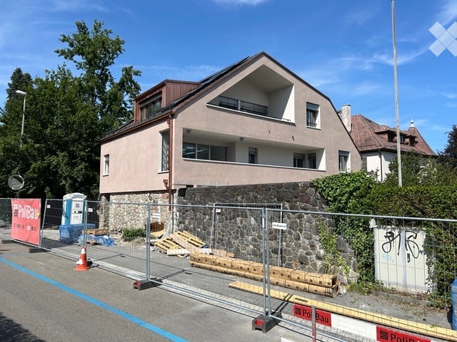 Two-story residential building with a modern design, featuring a stone base and a wooden upper section. The building has multiple windows and a balcony on the upper floor. The surrounding area includes a temporary fence and construction materials, indicat