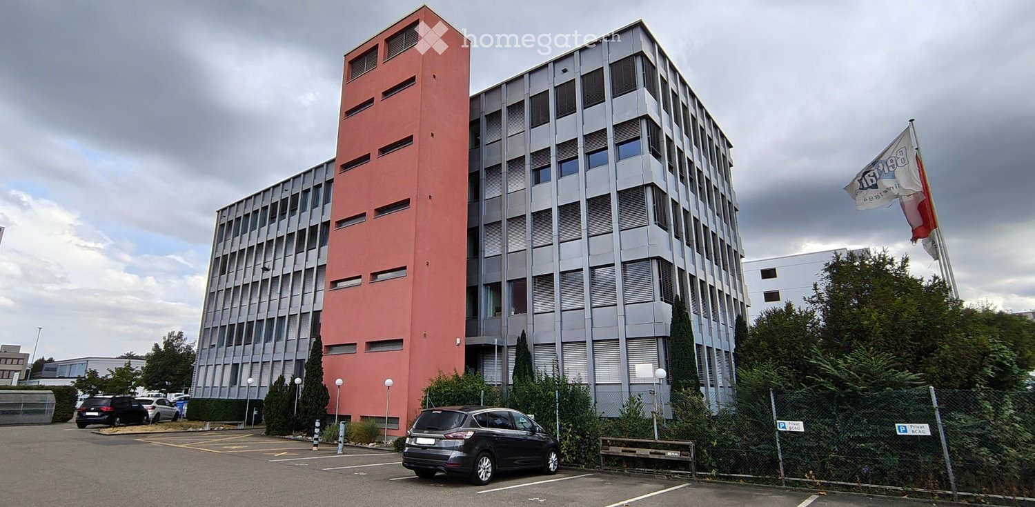 Modern multi-story building with a combination of red and grey facades, large windows, and a parking area in front.