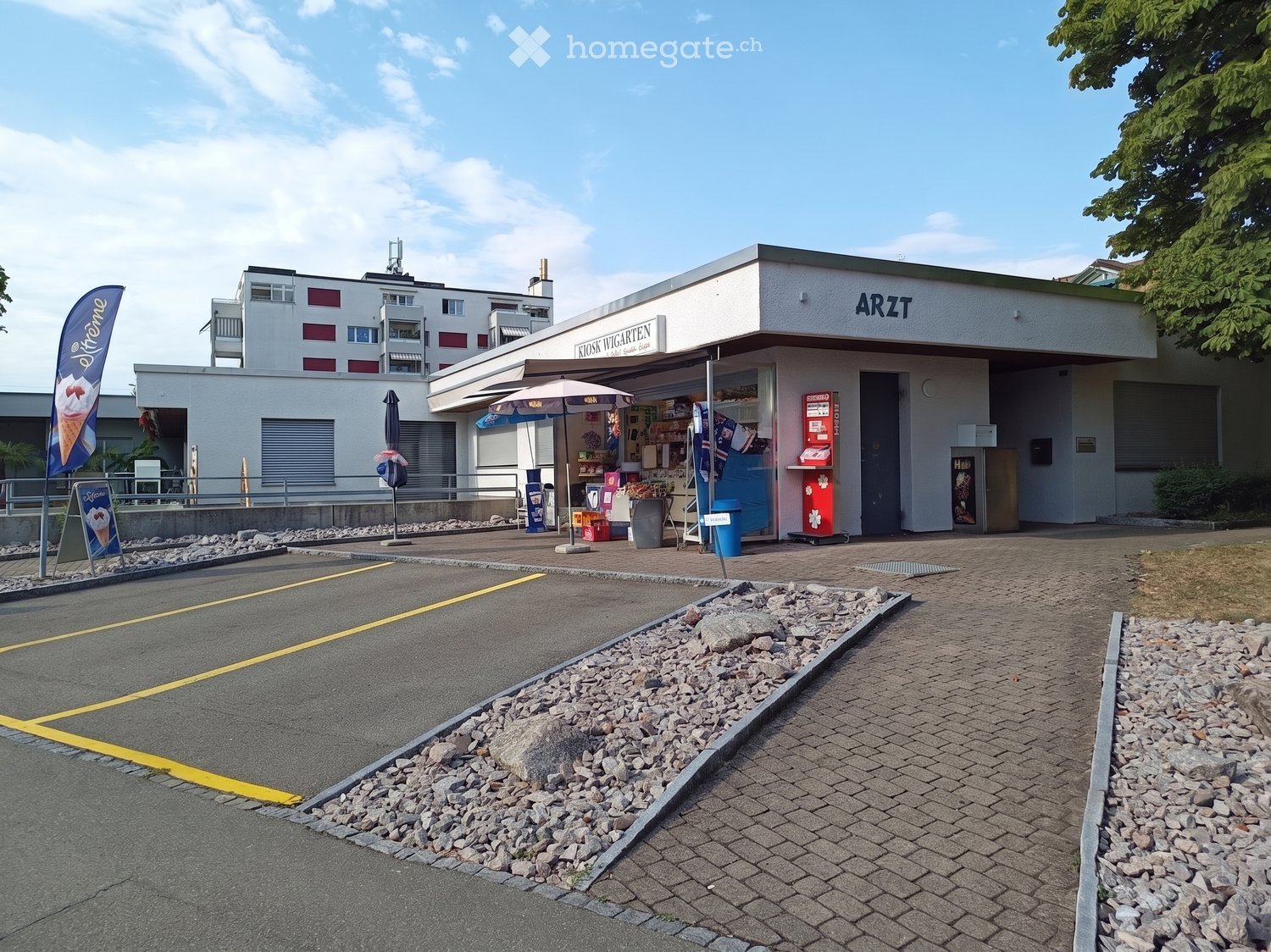 A small, modern building labeled "ARZT" with a kiosk area, umbrella, and vending machine visible. The surrounding area includes a paved walkway, parking space, and a multi-story residential building in the background.