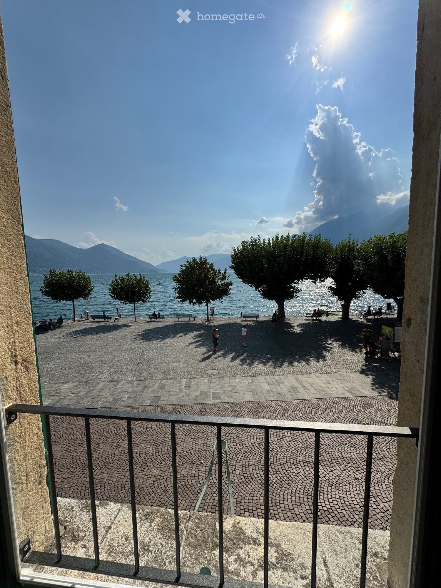 Balcony overlooking a scenic waterfront area with trees, benches, and distant mountains under a clear blue sky.