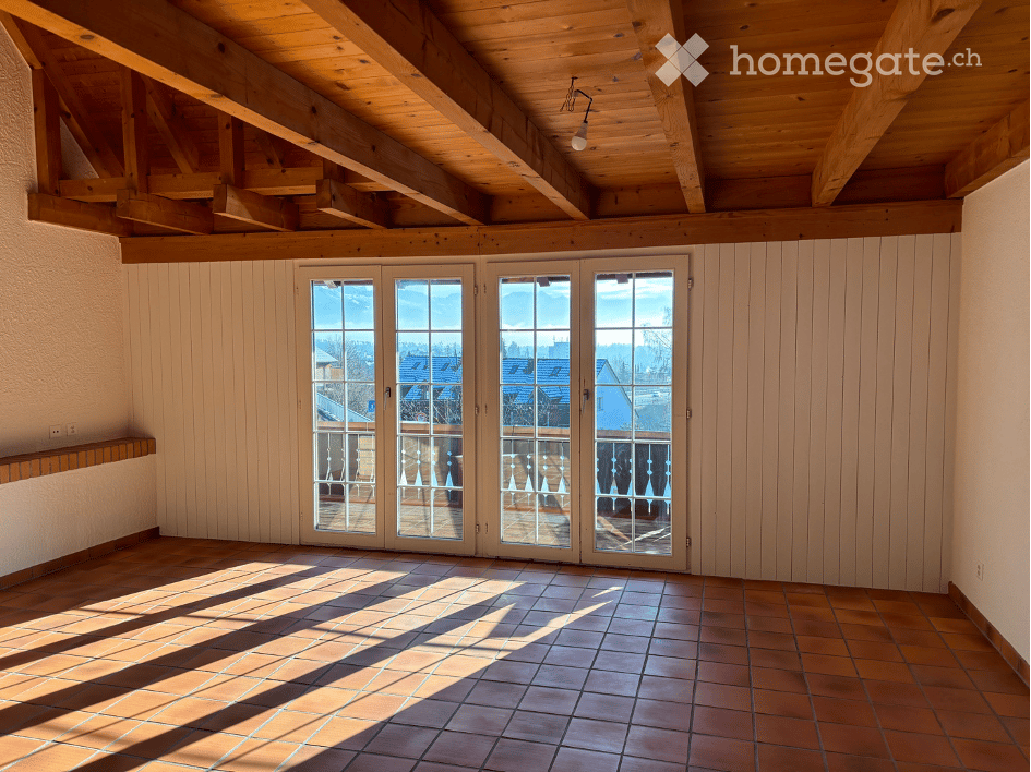 Empty room with exposed wooden beams on the ceiling, large glass doors leading to a balcony with a scenic view, tiled floor, and white paneled walls.
