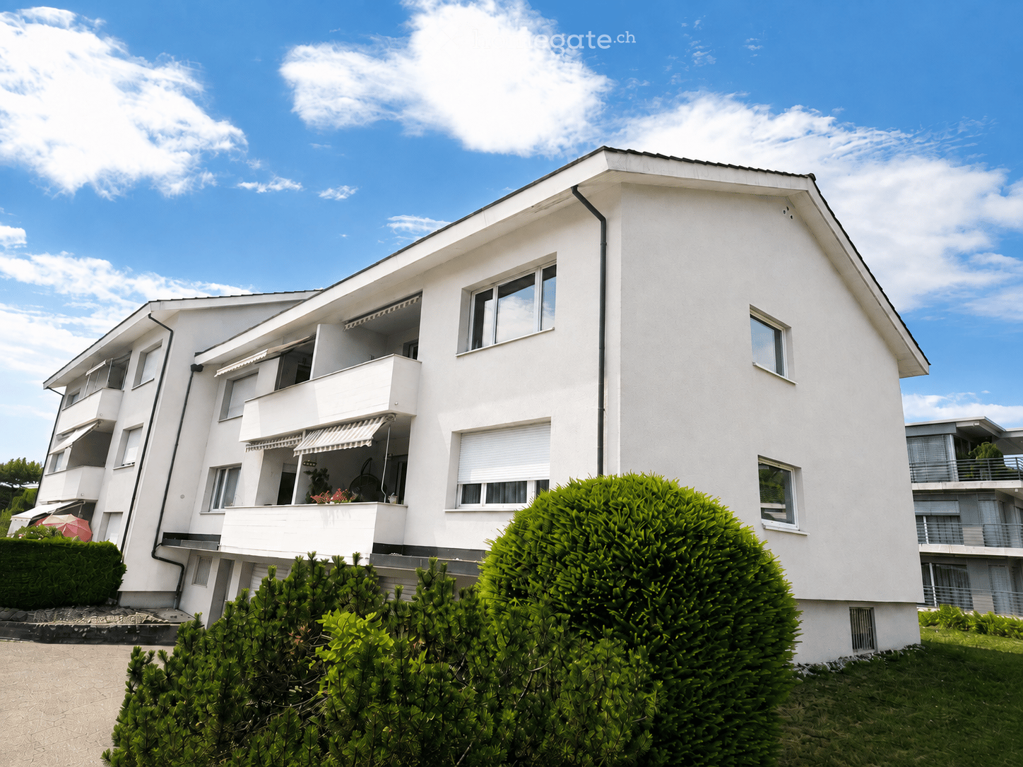 Modern white multi-story residential building with balconies, large windows, and neatly trimmed greenery in front.