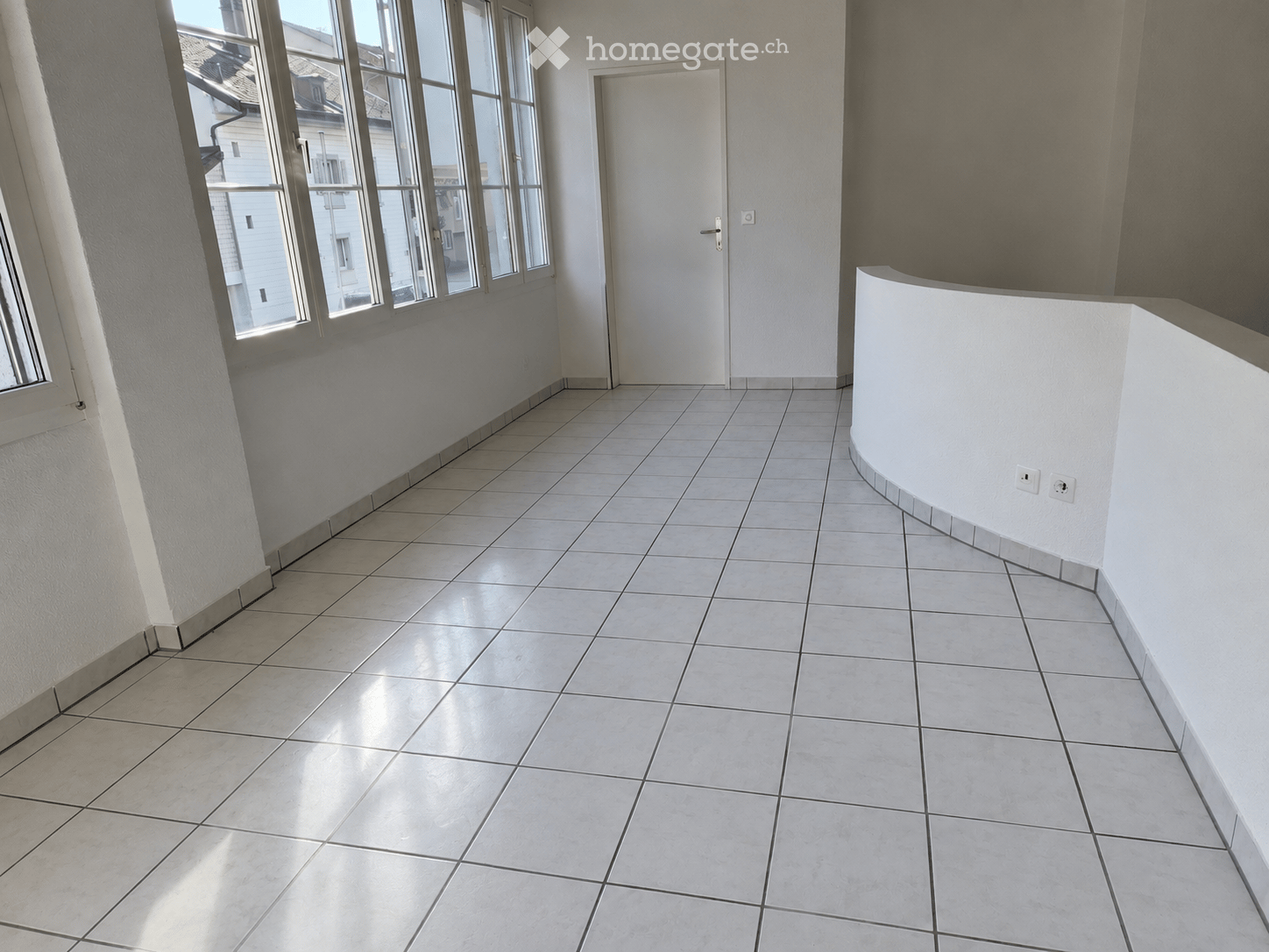 Empty room with tiled floor, white walls, large windows, and a white door.