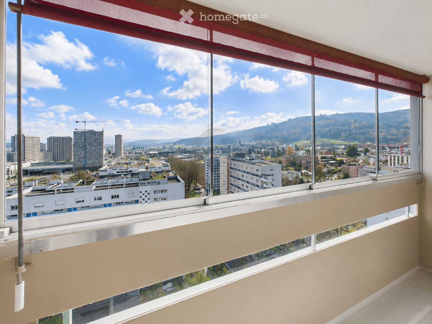 Large window with red blinds, offering a panoramic view of a cityscape and surrounding hills.