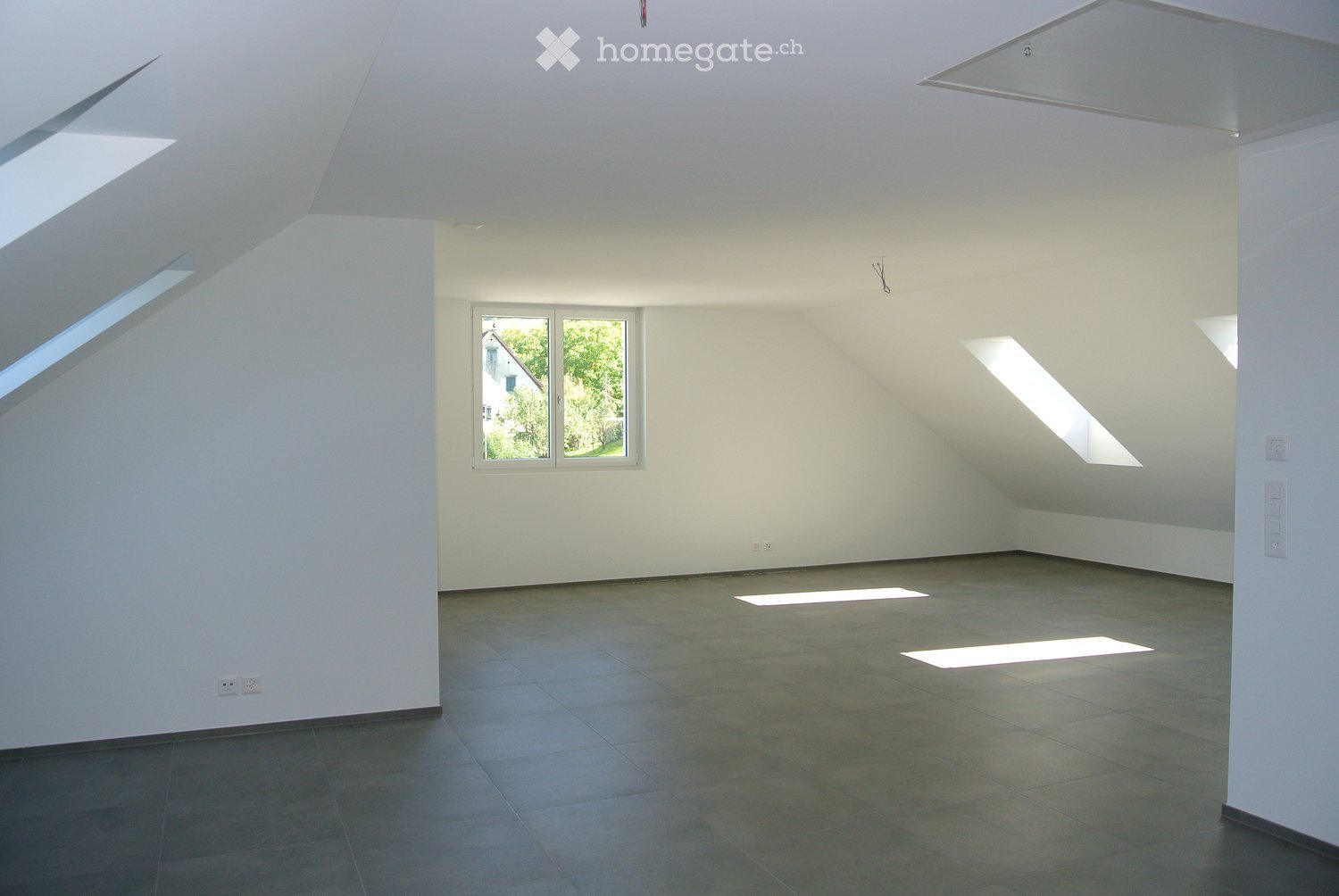 Empty room with white walls, tiled floor, and large windows allowing natural light to enter. The room has a high, slanted ceiling typical of an attic space.