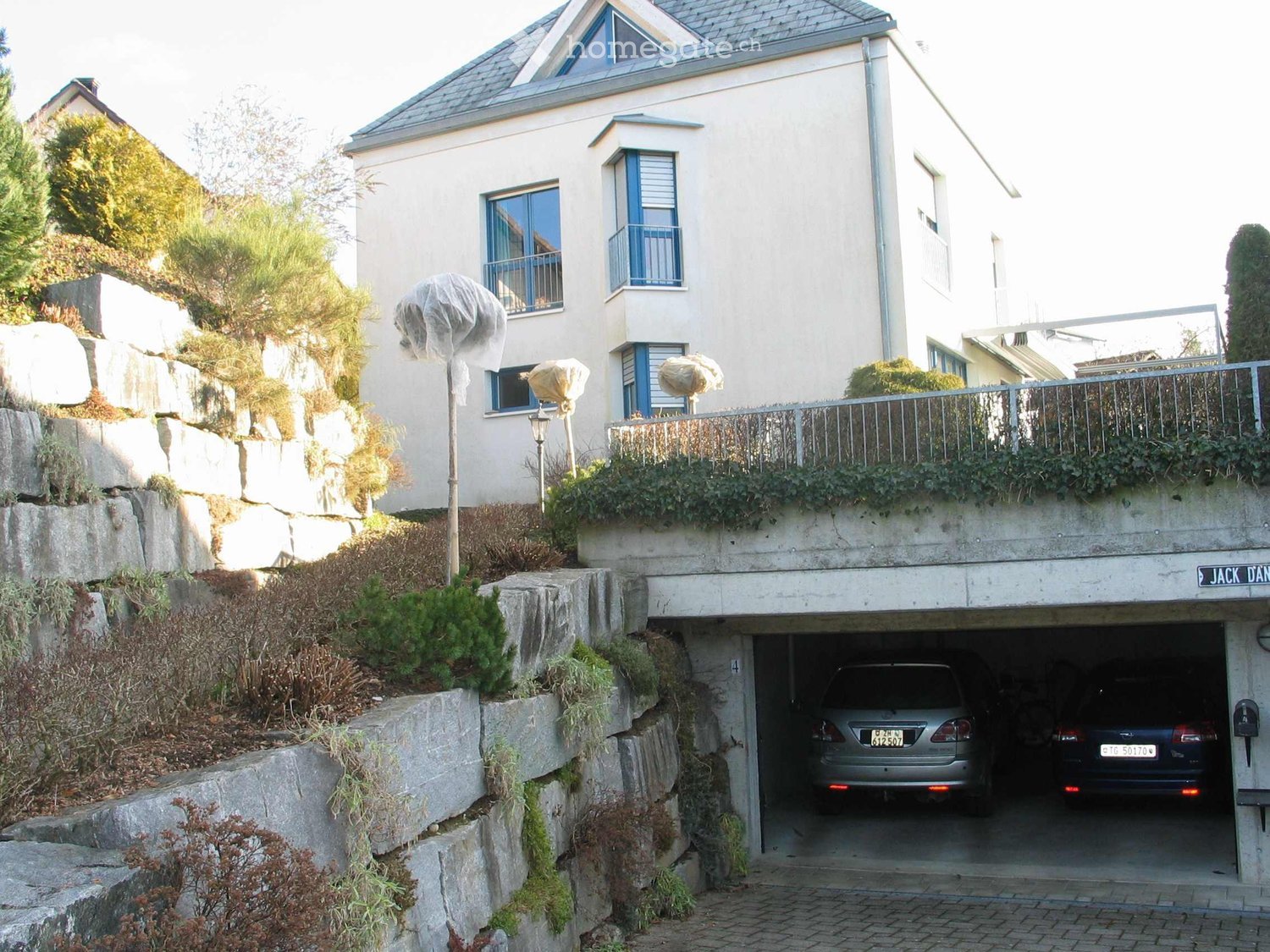 A two-story house with a garage, raised ground floor, and balcony. The garage has two parked cars, and the house is surrounded by greenery and stone walls.