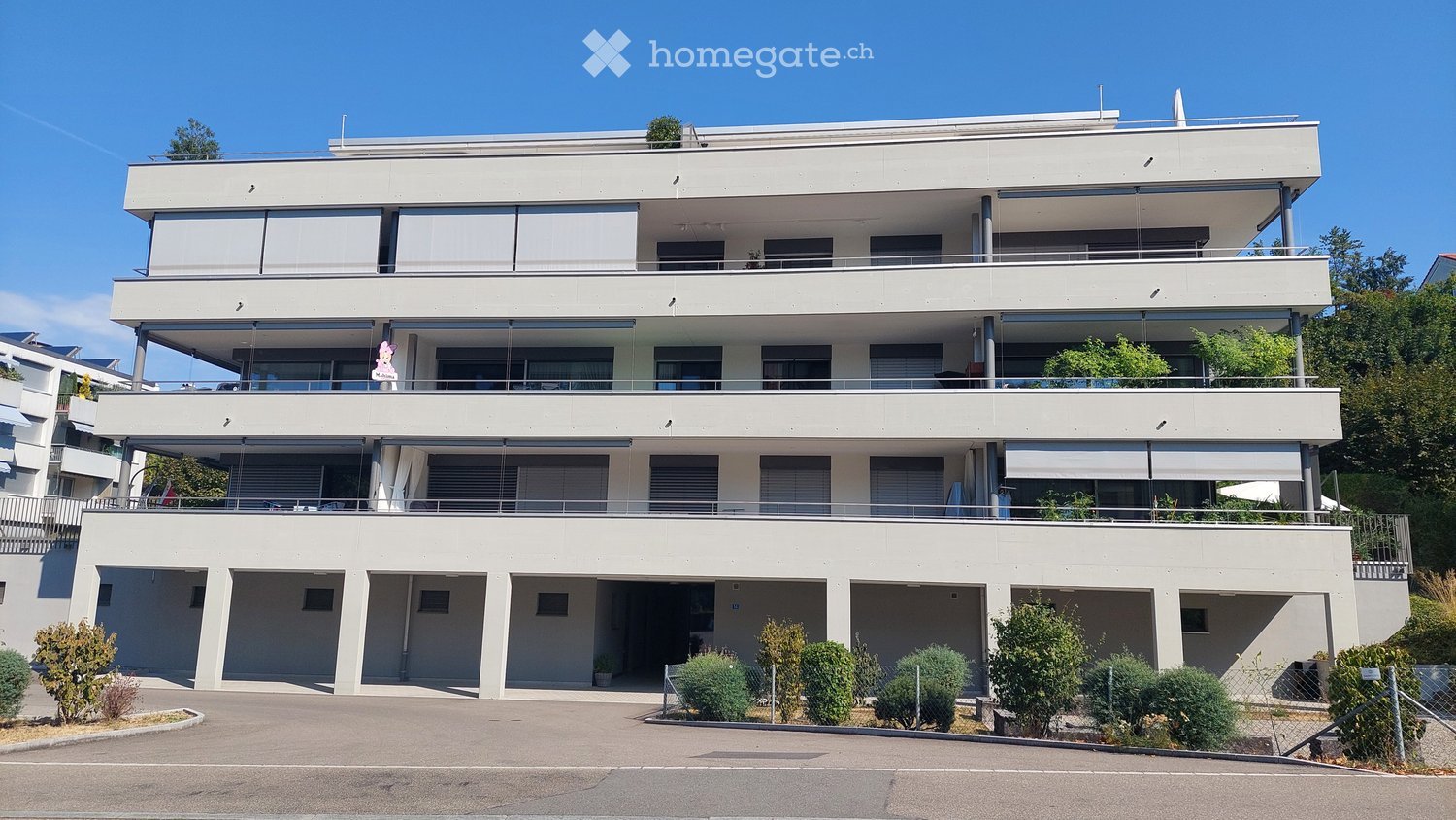 Modern three-story residential building with white facade, large windows, balconies, and a spacious parking area.
