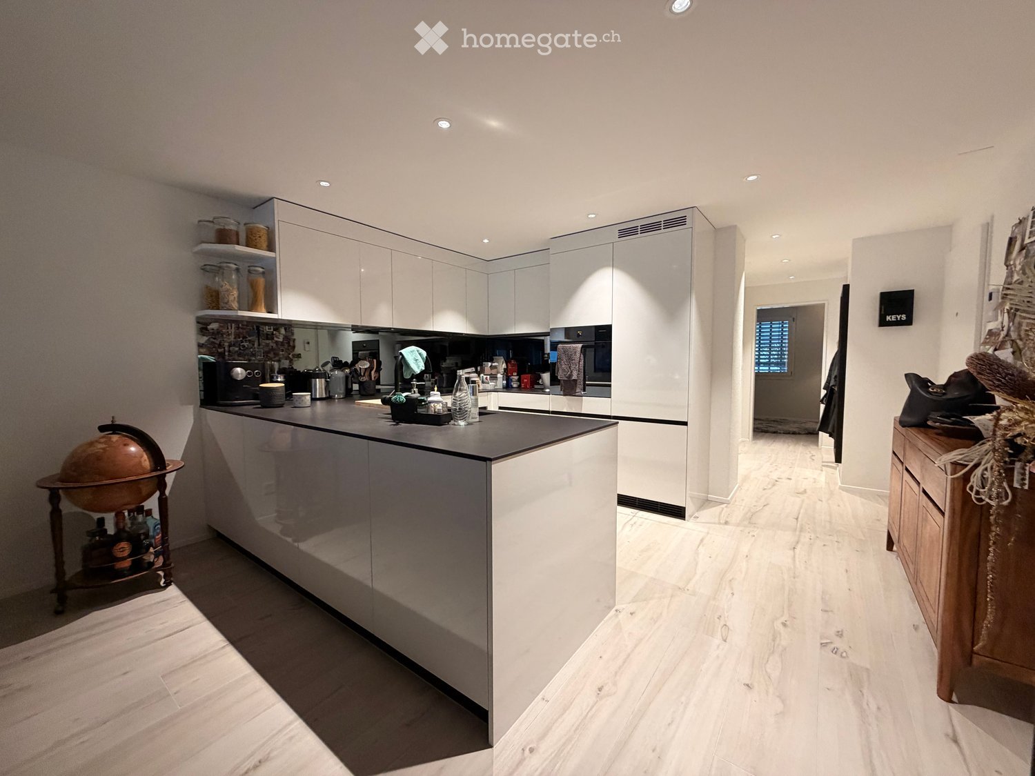 Modern kitchen with white cabinets, a central island, and various appliances including a coffee maker, fridge, and globe on a stand. The floor is light-colored wood, and the space is well-lit with recessed lighting.