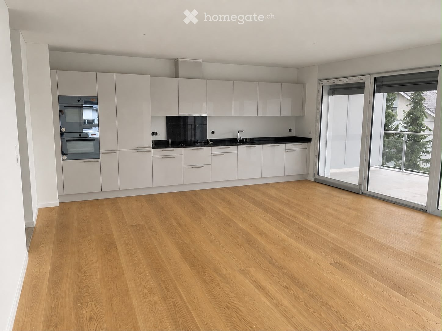 Empty kitchen with white cabinets, black countertops, and a large sliding glass door leading to a balcony with a view of trees.