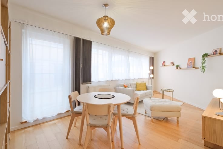 Bright living room with wooden flooring, large windows with white curtains, a sofa with yellow throw pillows, an ottoman, a dining table with chairs, and a hanging pendant light.