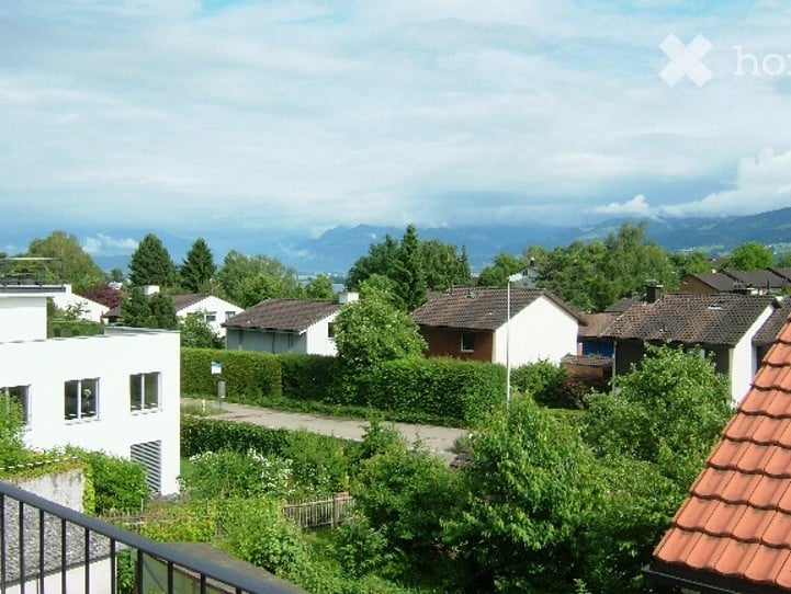 View from a balcony or terrace showing a suburban neighborhood with houses, trees, and distant mountains under a partly cloudy sky.
