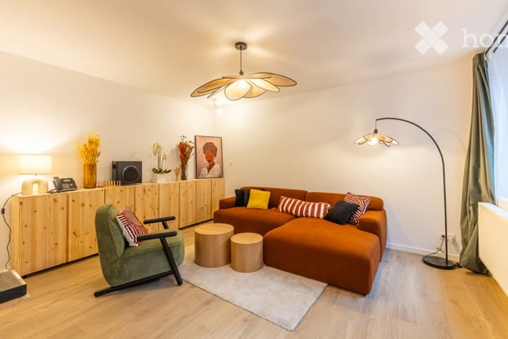 Modern living room with a large orange sectional sofa, green armchair, wooden coffee tables, floor lamp, and decorative vases on a wooden cabinet.