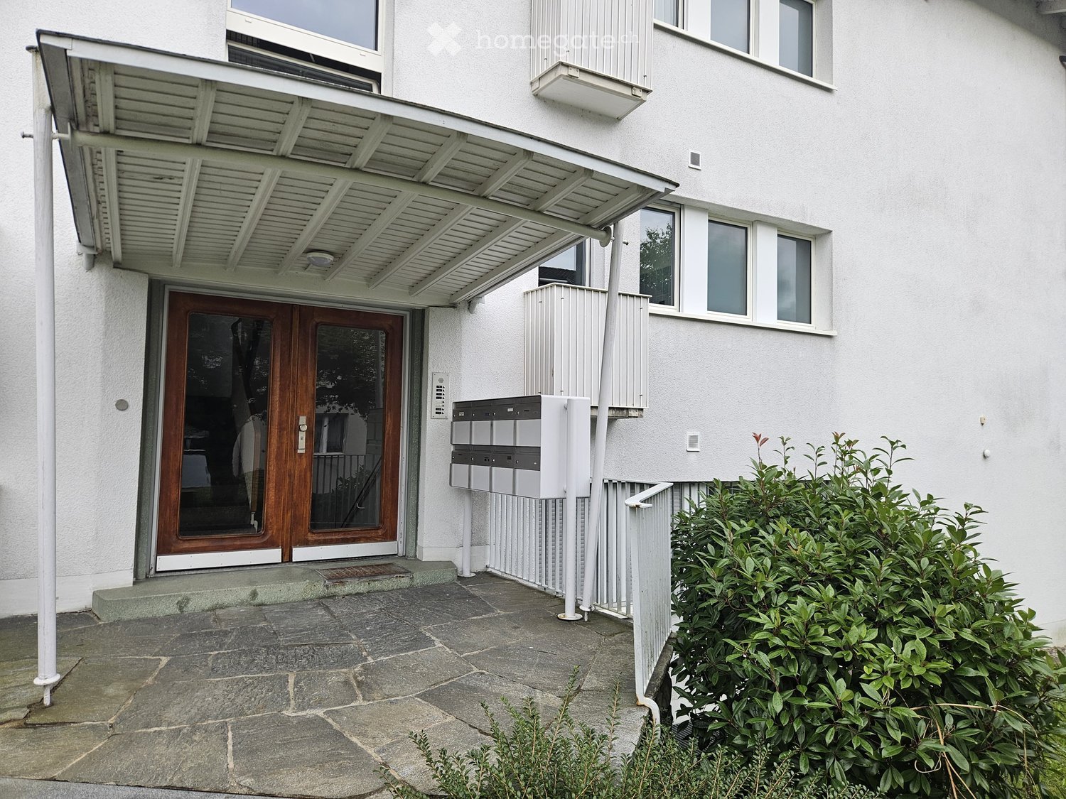 Modern building entrance with a covered porch, wooden double doors, mailboxes, and small garden area.