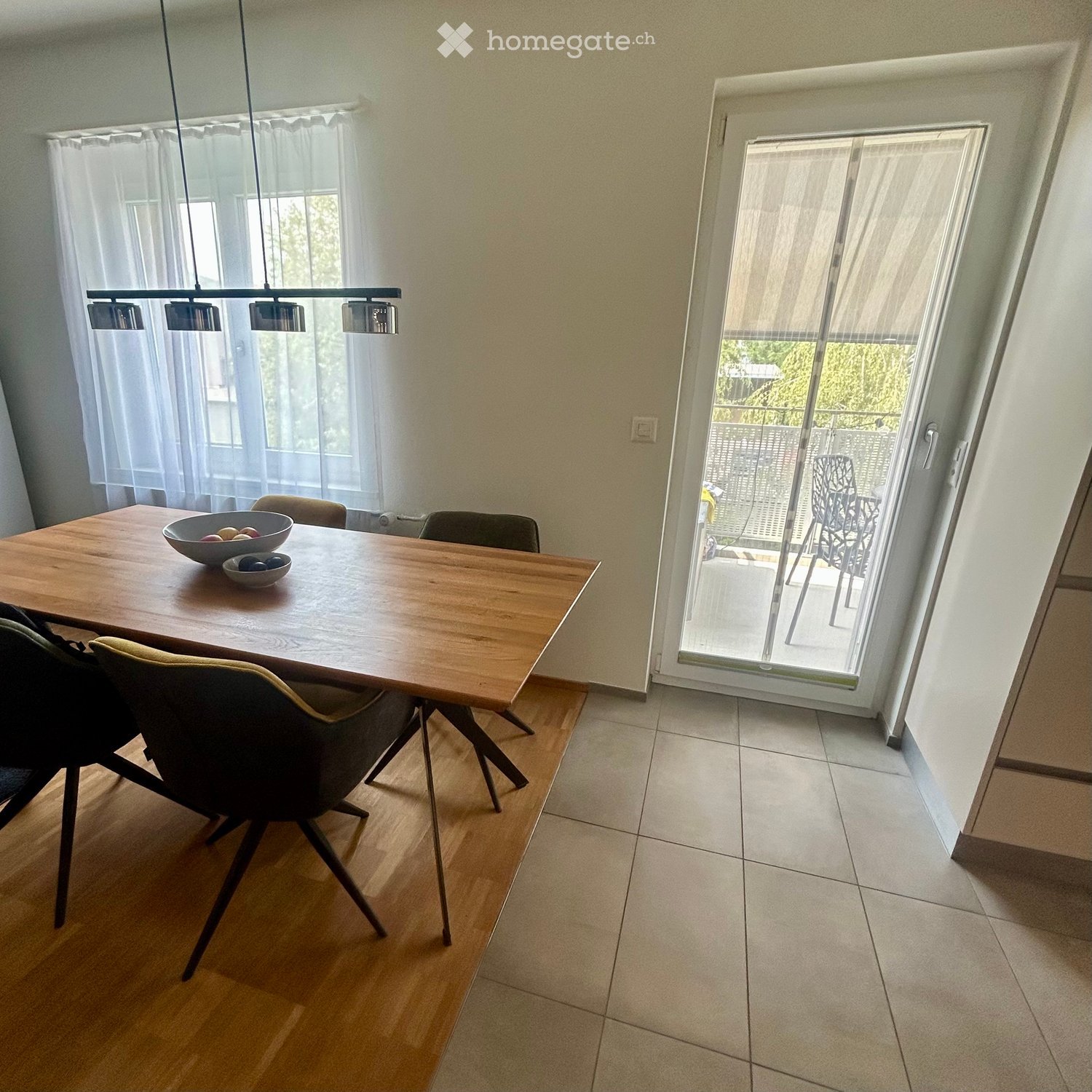 Dining room with a wooden dining table, chairs, and bowls on the table. Large windows with sheer curtains and a sliding glass door leading to a balcony.
