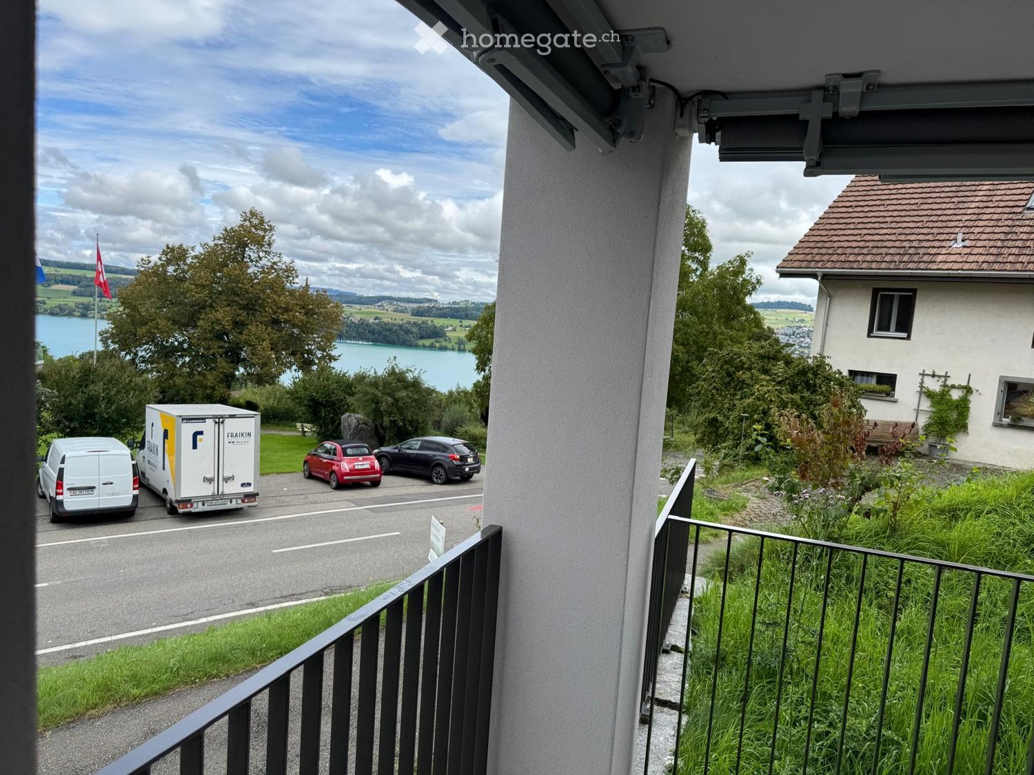 Balcony overlooking a street with parked cars, a moving van, and a scenic view of a lake and surrounding landscape.
