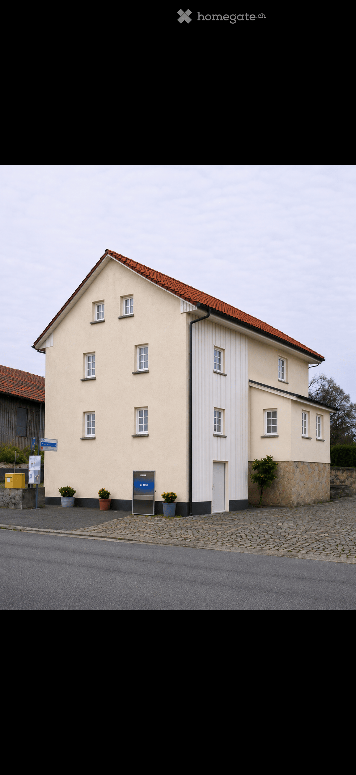 Two-story building with beige walls, red-tiled roof, multiple windows, and a white door. Potted plants and an alarm box are visible near the entrance.