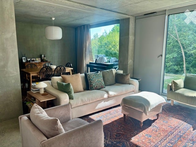 Modern living room with large windows offering a view of greenery, featuring a cream-colored sofa, ottoman, armchair, coffee table, dining table, and chairs.