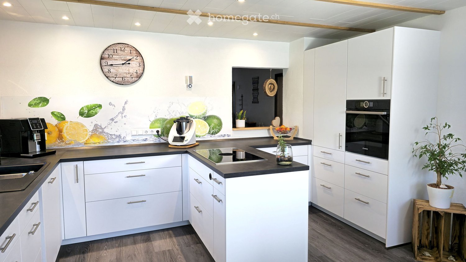 Modern kitchen with white cabinets, black countertops, coffee machine, blender, oven, fruit bowl, and a small plant on a wooden stool.