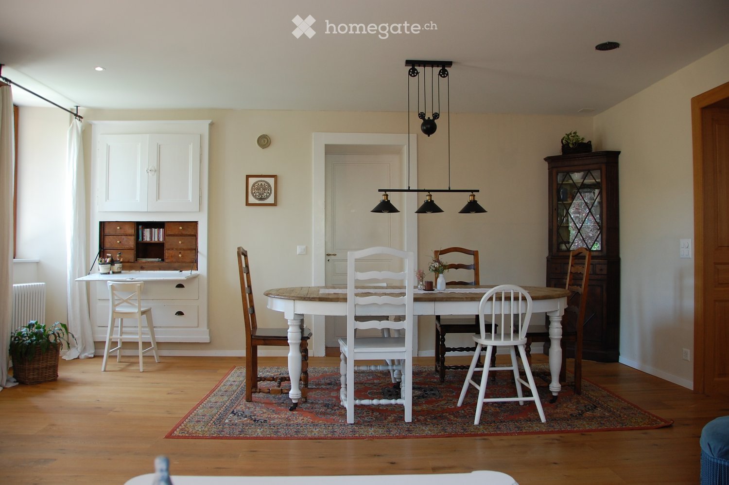 Bright dining area with wooden floor, large wooden dining table surrounded by white and wooden chairs, a hanging light fixture, a white cabinet with drawers, a small desk with a stool, and a dark wooden cabinet with glass doors.