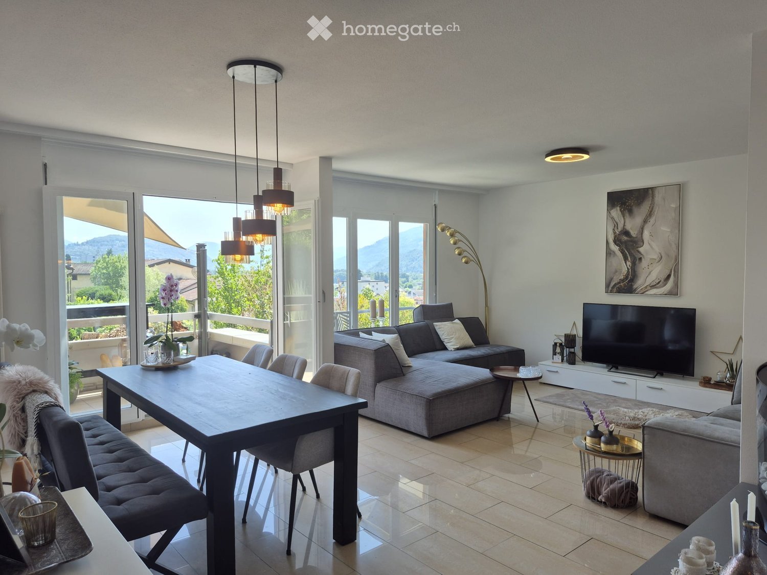 Modern living room with large windows offering a scenic view, featuring a grey sectional sofa, coffee table, dining table with chairs, floor lamp, and a television on a stand.