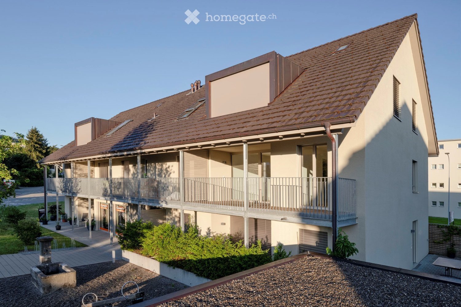 Modern two-story building with a brown tiled roof, white walls, and a balcony on the upper floor. The building features a small garden area with shrubs and a paved walkway.