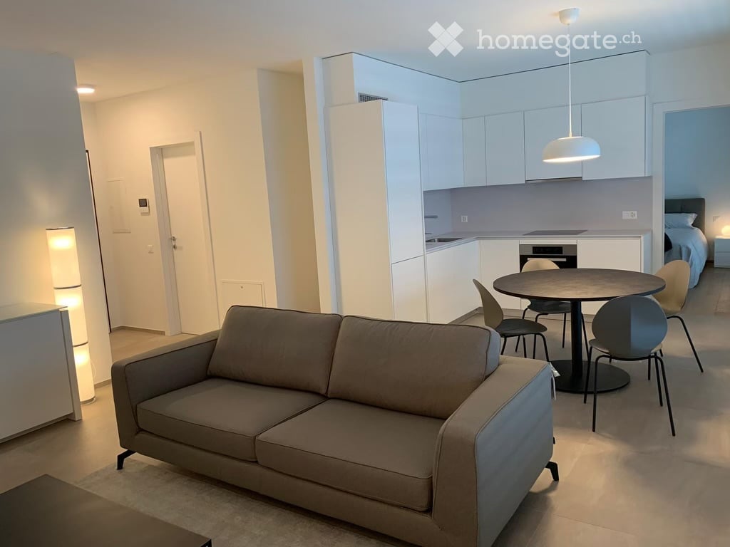Modern, open-plan living and dining area with a sofa, dining table, chairs, and a small kitchen area with cabinets and a bed visible in the background.