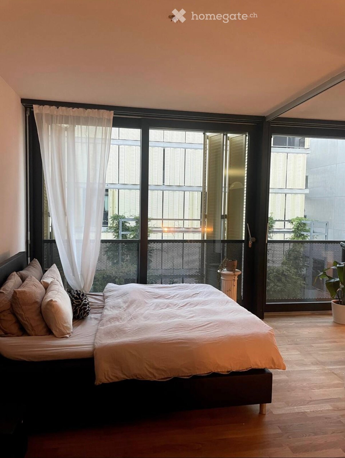 Modern bedroom with a large bed, multiple pillows, sheer white curtains, and a view of a balcony with plants.