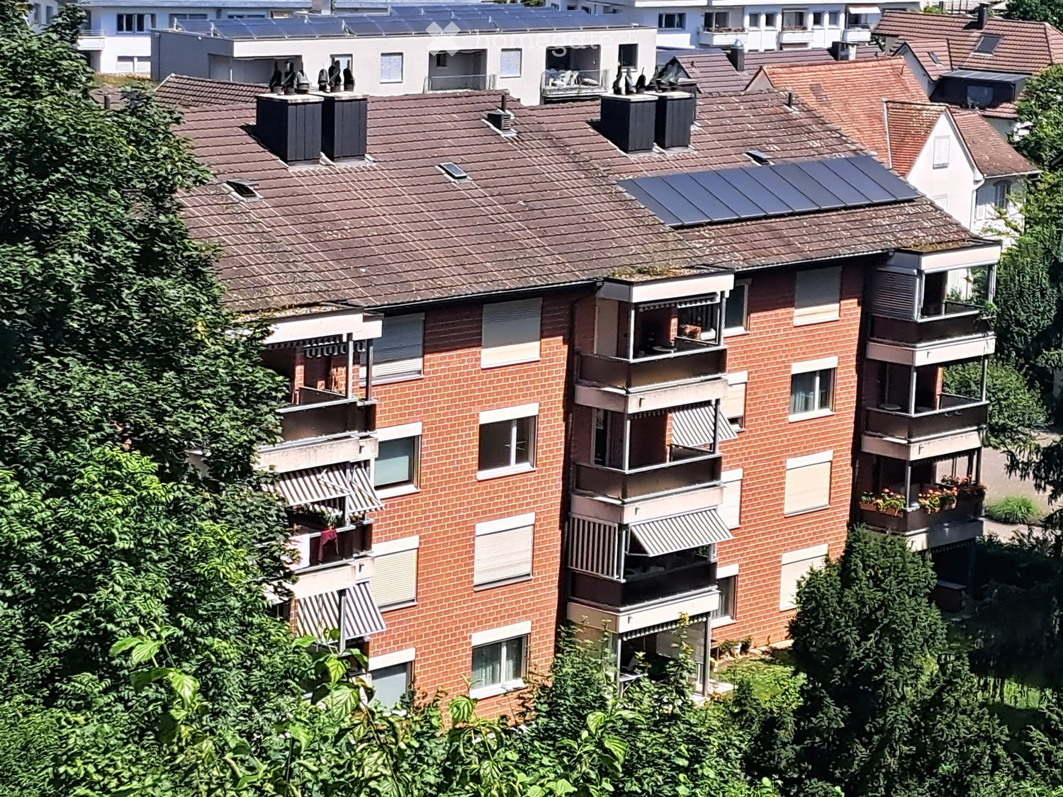 Multi-story brick building with multiple balconies, solar panels on the roof, and surrounding greenery.