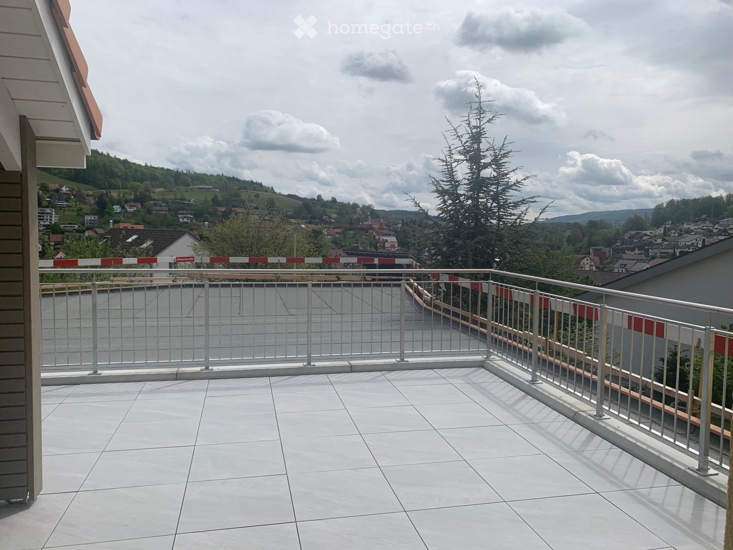 Balcony with white tiled floor, metal railing, and scenic view of a town and hills under a partly cloudy sky.