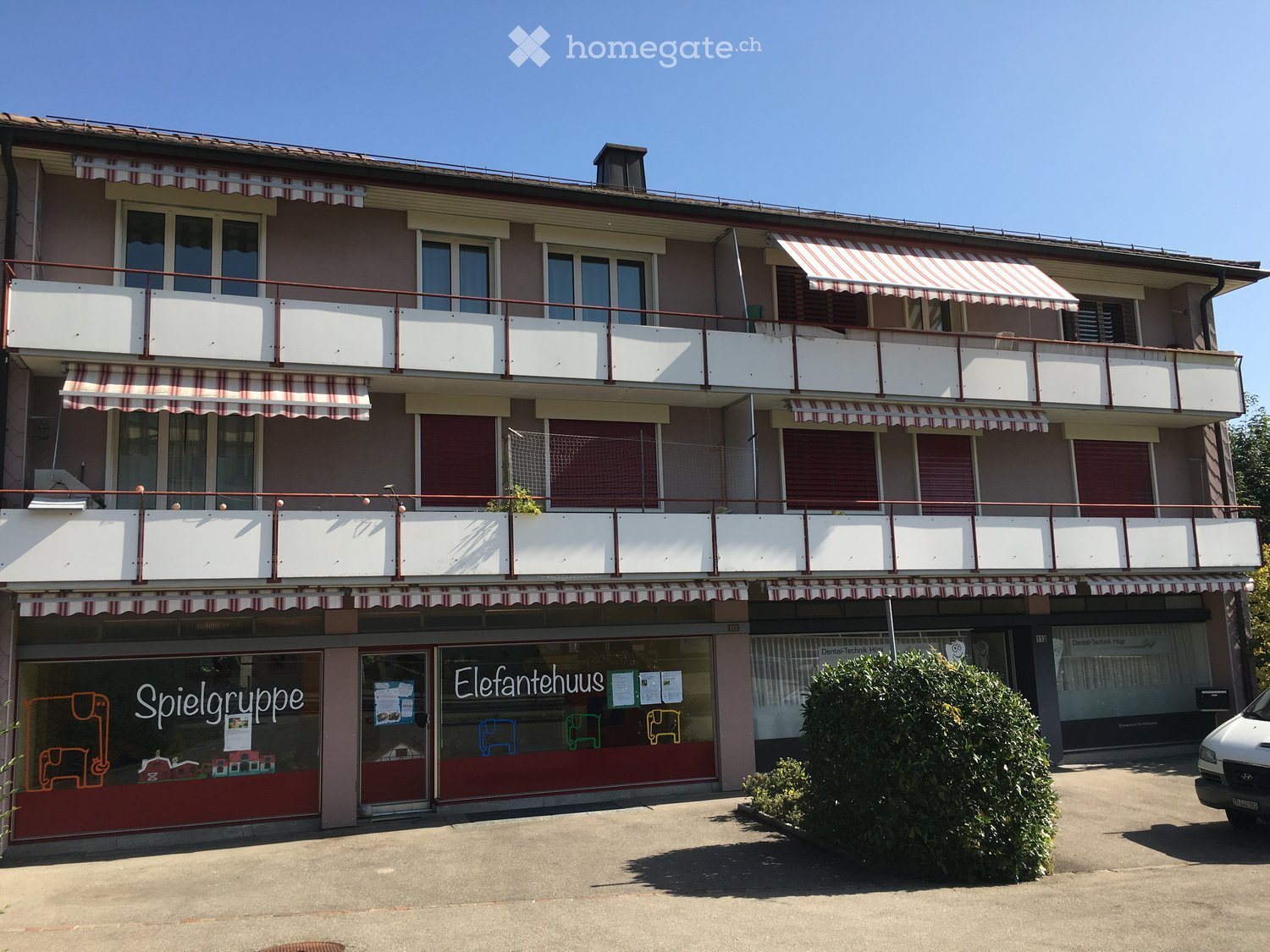 Three-story building with balconies, striped awnings, and storefronts including 'Spielgruppe' and 'Elefantenhaus'.
