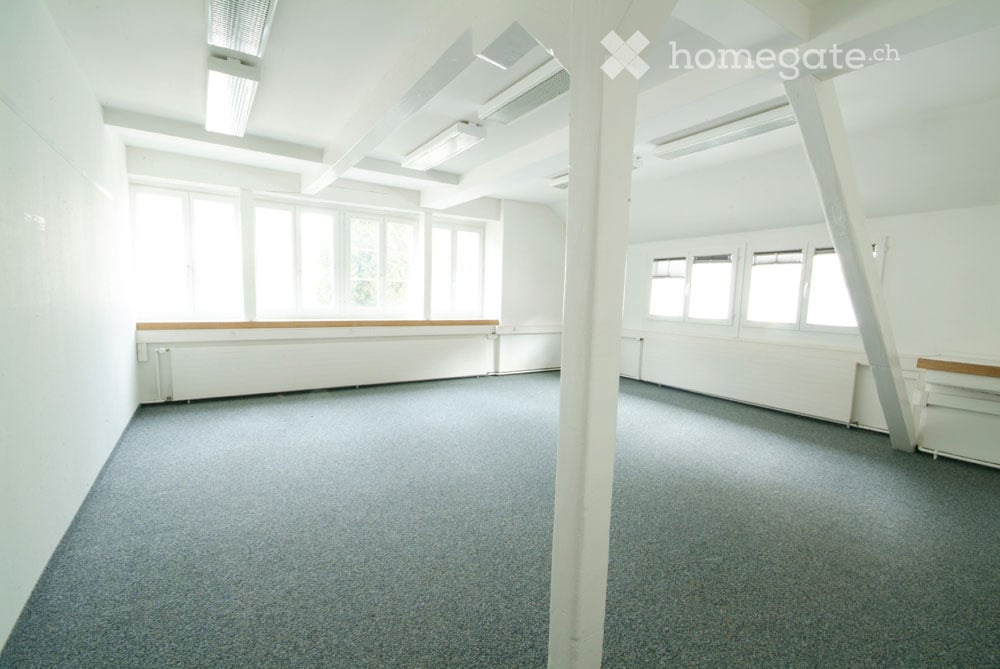 Empty room with white walls, grey carpet, large windows, and exposed ceiling beams.