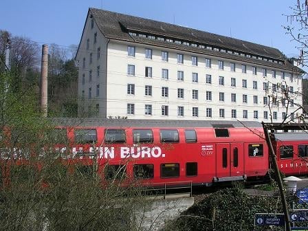 A large, multi-story building with white walls and numerous windows, situated near a railway track with a red train passing by.