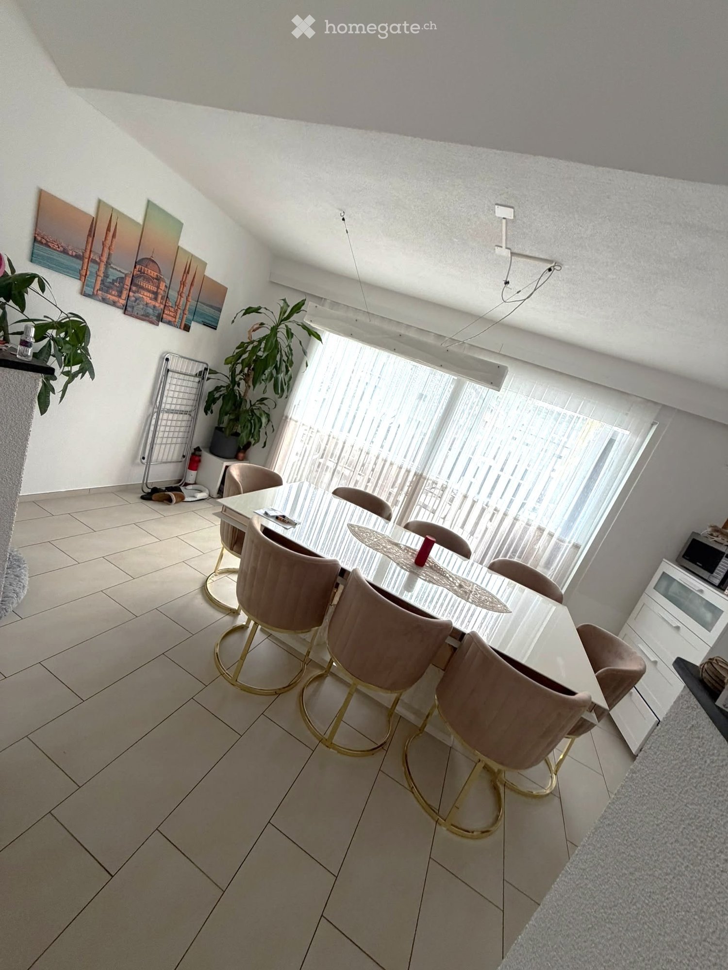 Dining area with a modern glass dining table, six beige chairs with gold bases, large windows with sheer curtains, and a potted plant near the window.