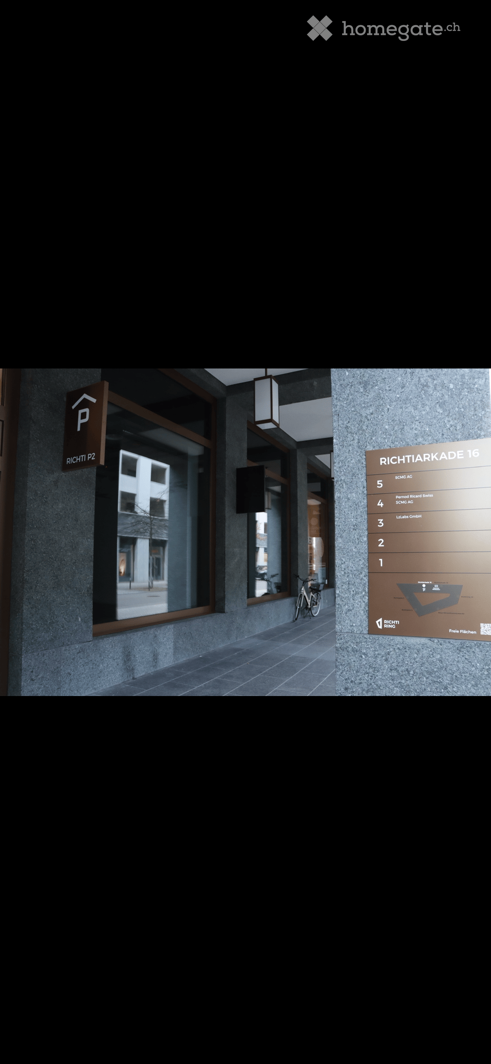 Modern hallway with a signboard listing office numbers, a bicycle parked nearby, and a view of a building through a large window.