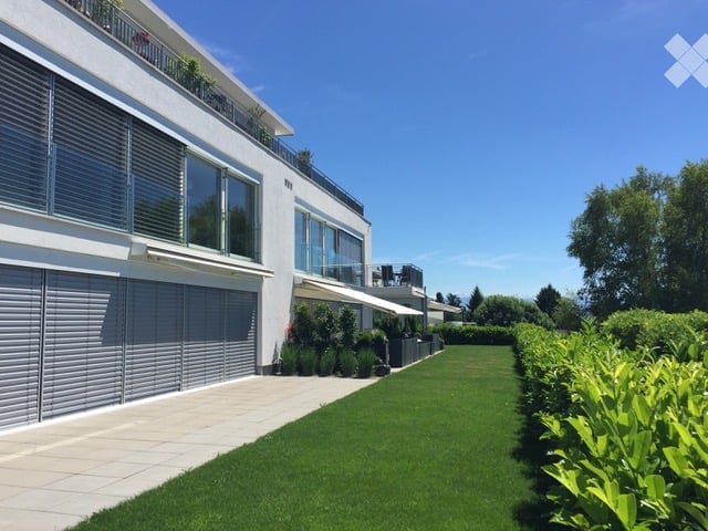 Modern white building with large windows, balconies, and a well-maintained lawn and hedge border.