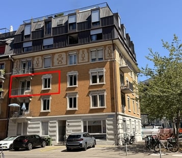 Multi-story apartment building with a mix of brick and plaster facades, multiple windows, and balconies. The ground floor has a storefront with a glass entrance. Several cars are parked in front, and bicycles are lined up on the right side.