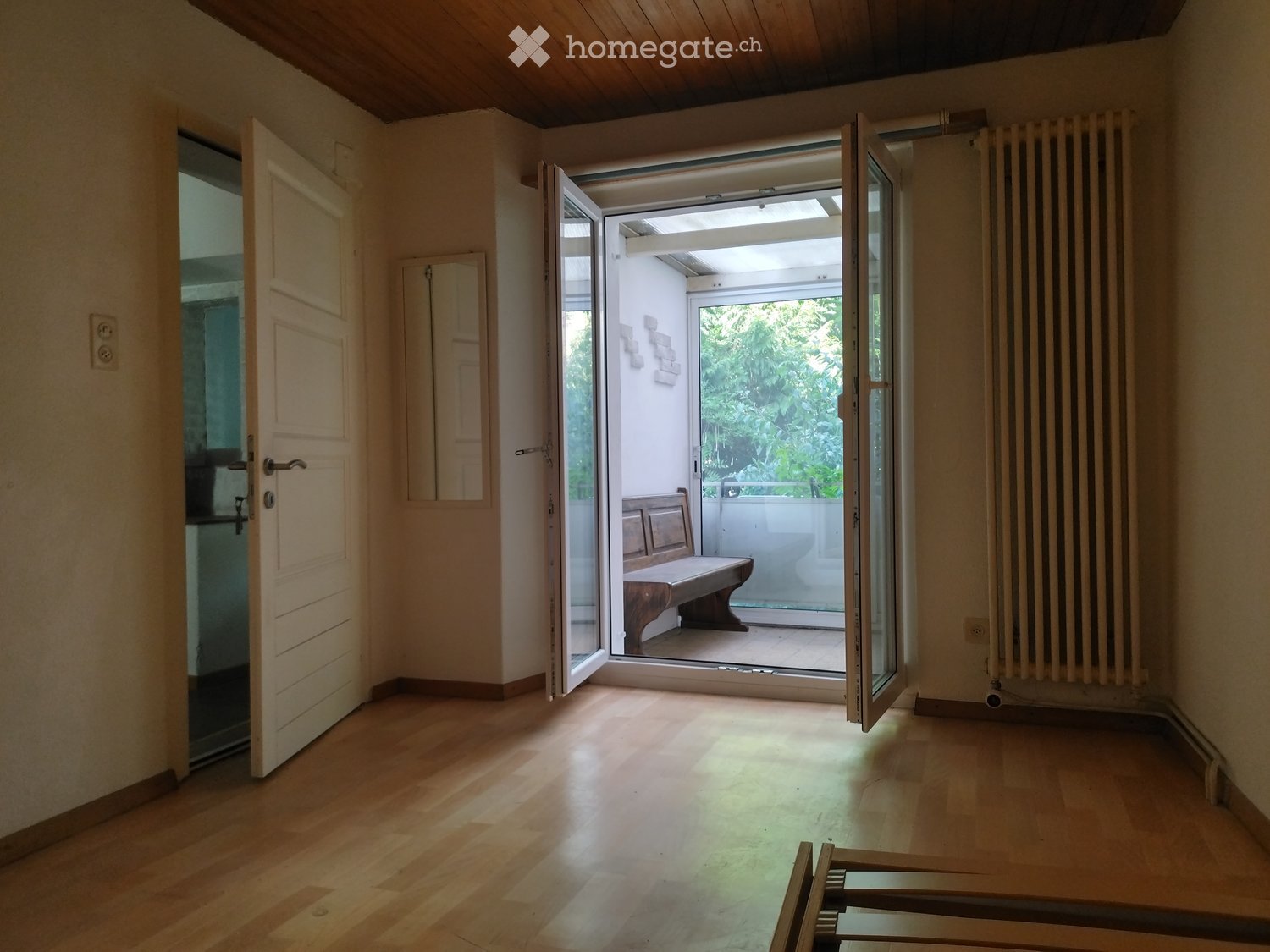 Empty room with wooden flooring, large open sliding doors leading to a balcony area, radiator on one wall, and a visible bench outside.