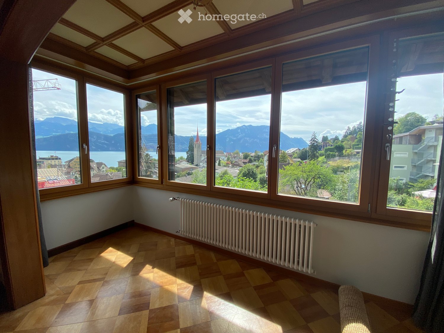 Empty room with wooden flooring, large windows offering a scenic mountain and lake view, and a white radiator along the wall.