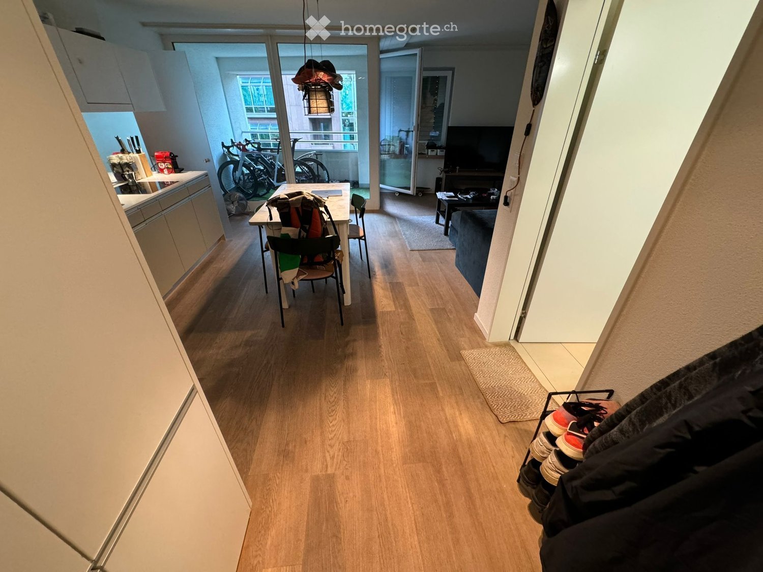 A well-lit hallway with wooden flooring, leading to a kitchen and living area. The hallway features a shoe rack on the right and a small rug near the entrance.