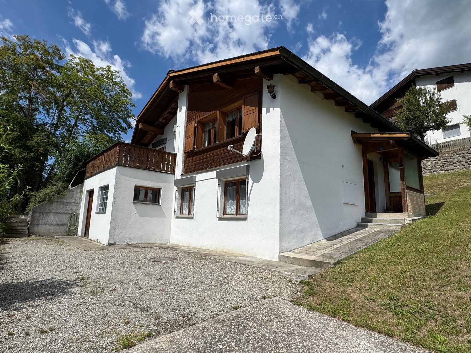 Two-story white house with wooden accents, satellite dish, and balcony on the upper level. The house is surrounded by greenery and has a gravel driveway.