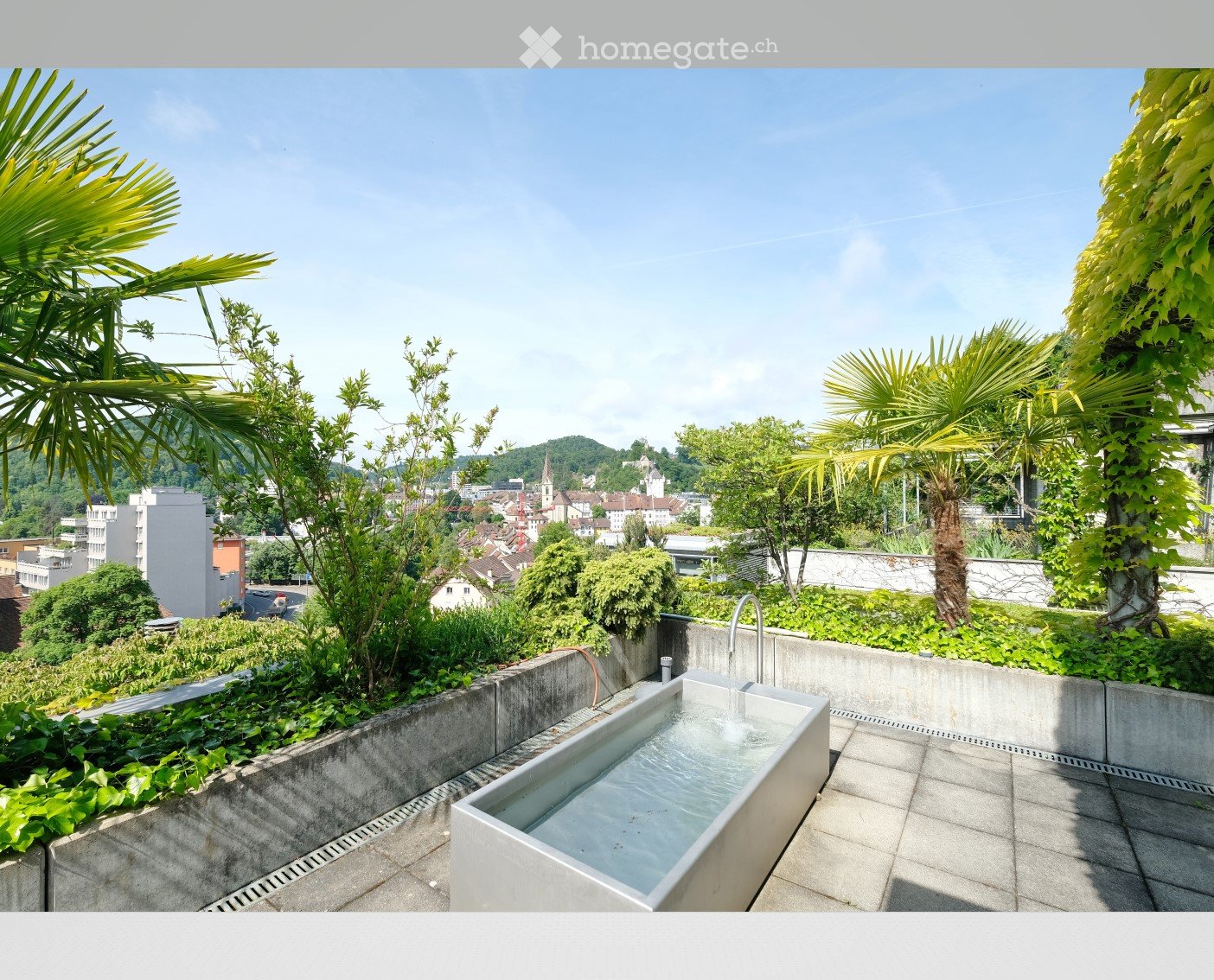 A modern rooftop terrace with a minimalist concrete bathtub, surrounded by lush greenery and palm trees, offering a scenic city view.