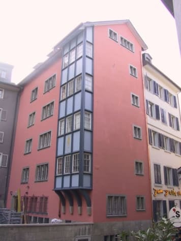 Multi-story residential building with pink facade and blue-framed windows, featuring a balcony on the third floor.