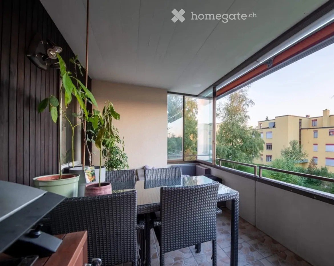 Balcony with a glass table, wicker chairs, and potted plants, offering a view of the city and greenery.