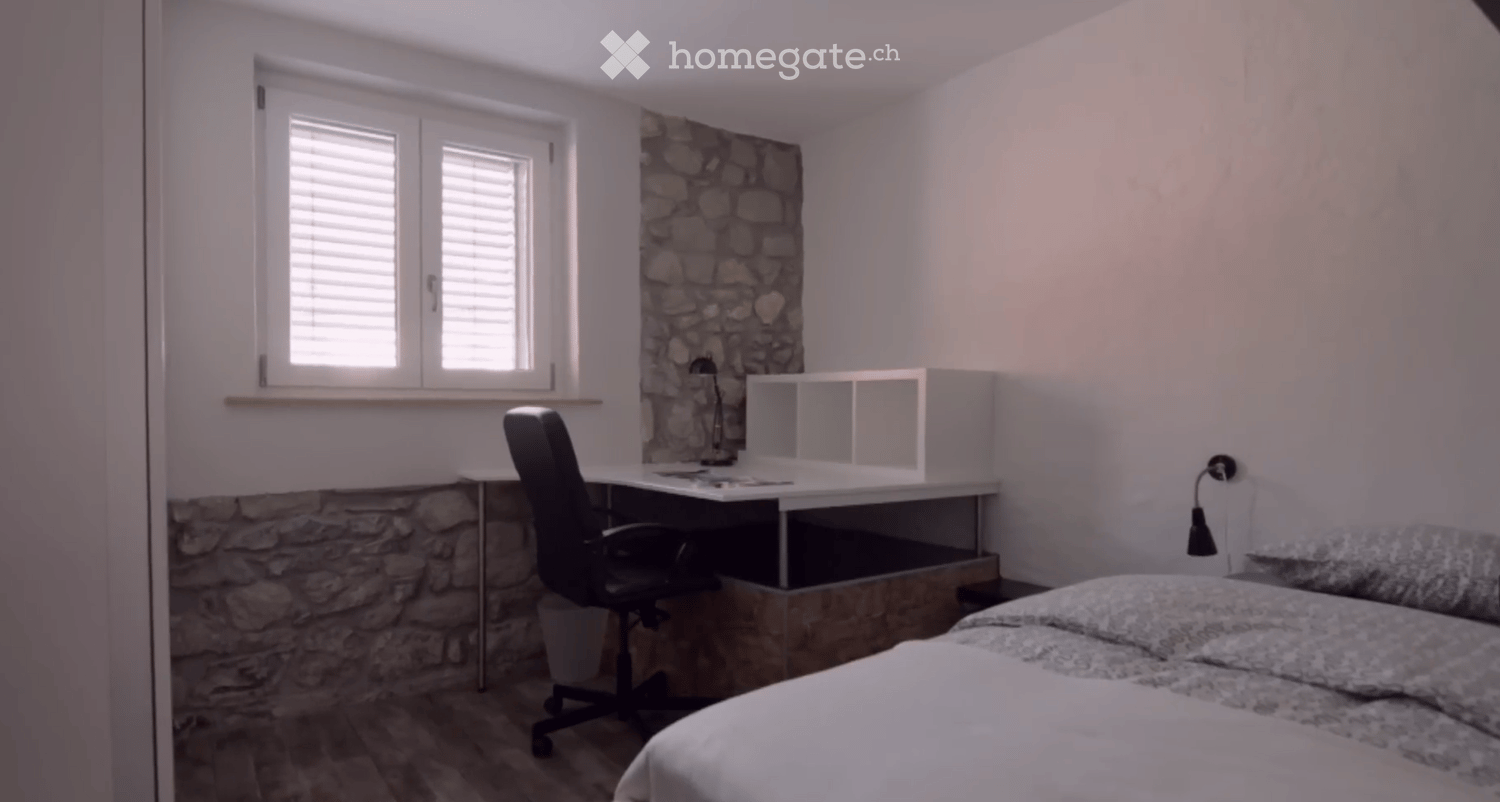 Cozy bedroom with a bed, desk, chair, and a stone accent wall. The room features a window with blinds and a modern storage unit.