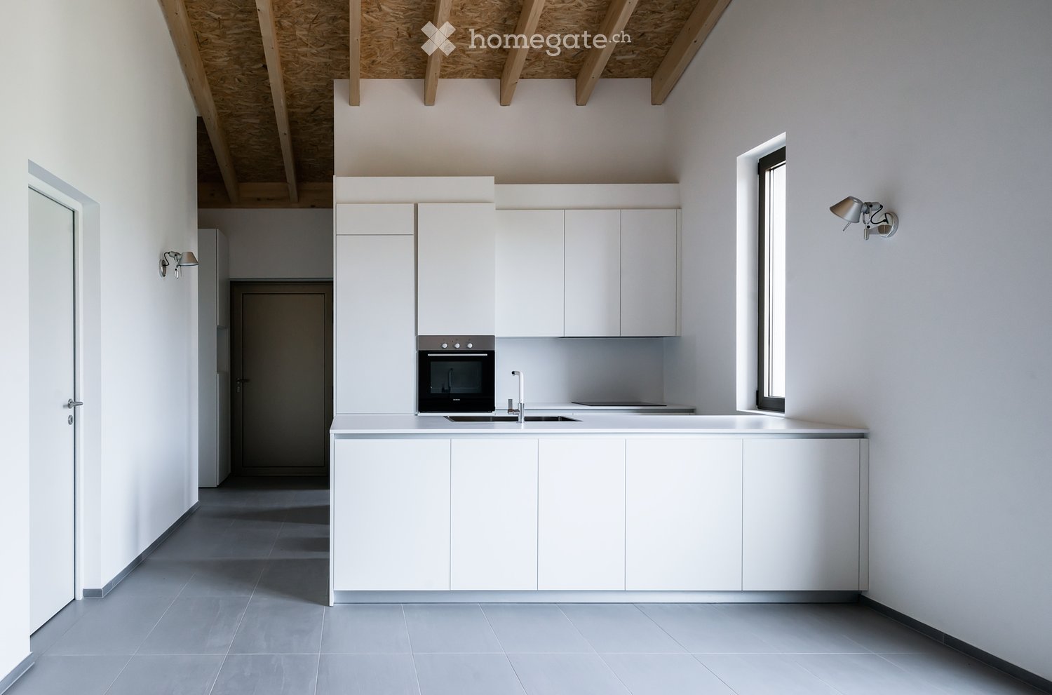 Modern, minimalist kitchen with white cabinets, stainless steel oven, sink, and faucet. Exposed wooden beams on the ceiling and a large window provide natural light.
